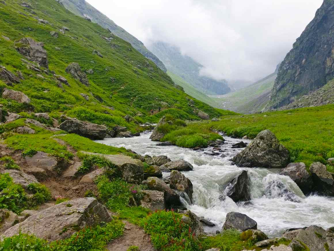 Supin river flowing through the valley in between huge mountains covered with green grass and grey fog filling the valley in back